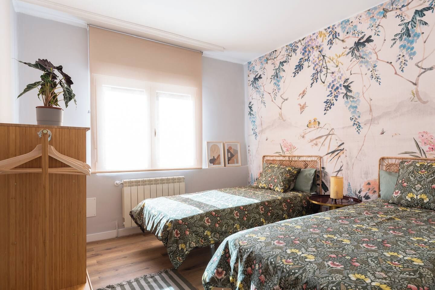 A bedroom with two beds, a radiator, a wooden wardrobe, a window with blinds, and a floral wallpaper.