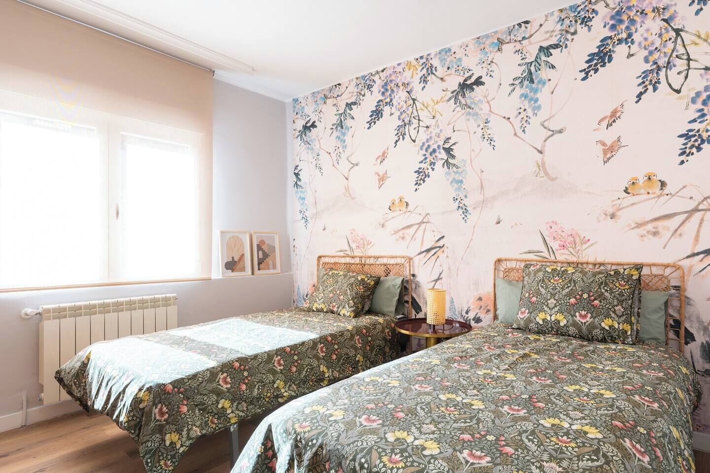 A room with two beds, a radiator, a window, and a wall with a floral mural.