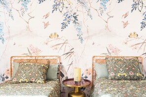 A room with two wicker sofas, patterned cushions, a round table with a lamp, and a floral wallpaper.