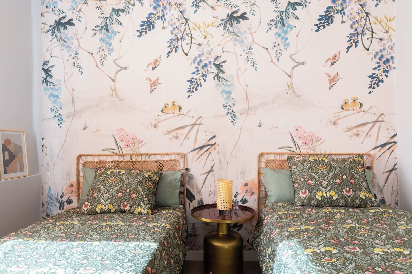 Une chambre avec deux lits, chacun agrémenté de draps à motifs floraux, une table ronde avec une lampe et un mur orné d’une fresque sur le thème de la nature.