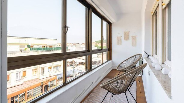 A balcony with large windows, wicker chairs, and a view of the cityscape. - Casa Surfer Somo centro (Somo)