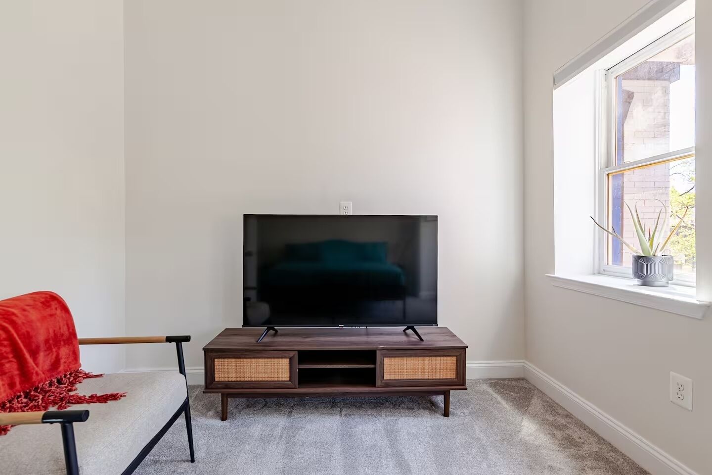 Una stanza con una TV a schermo piatto su un supporto in legno, una sedia rossa, una finestra con vista su un edificio e una pianta in vaso sul davanzale.