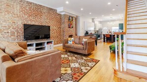 Ein Wohnzimmer mit Backsteinwand, einem Fernseher, Sofas und einer Küche im Hintergrund.
