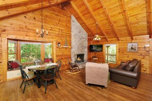 Un intérieur de chalet chaleureux avec une cheminée en pierre, des murs en bois, un coin repas et un espace détente avec un canapé et un téléviseur.