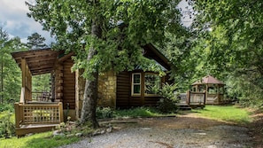 Un rustico chalet con veranda e gazebo, circondato da alberi fitti e un vialetto sterrato.