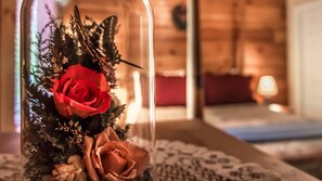 Un vase en verre contenant une rose rouge et une plus petite rose, posé sur une nappe à motifs dans une pièce aux murs en bois, éclairée d’une lumière chaleureuse.