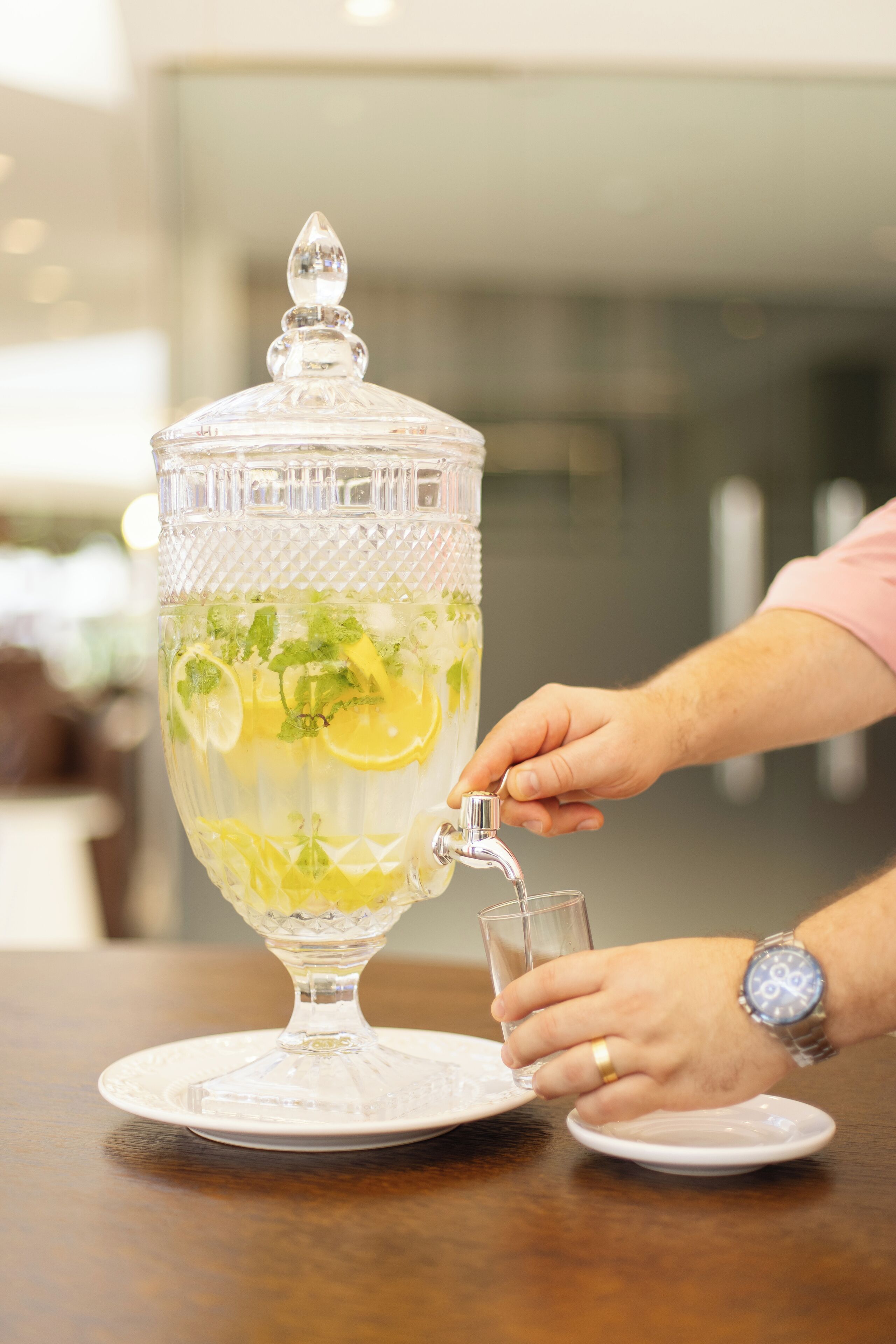 Una fuente de vidrio transparente llena de bebida, adornada con rodajas de limón y hojas de menta, sobre un plato blanco.