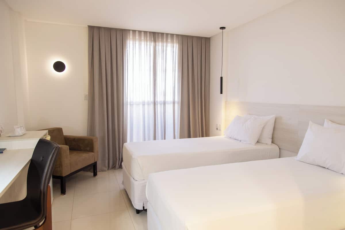 Une chambre d’hôtel avec deux lits, un bureau, une chaise et une grande fenêtre avec des rideaux.