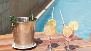 Escena junto a la piscina con un cubo metálico que contiene botellas, dos vasos con rodajas de limón y pajitas, y una mesa de madera.