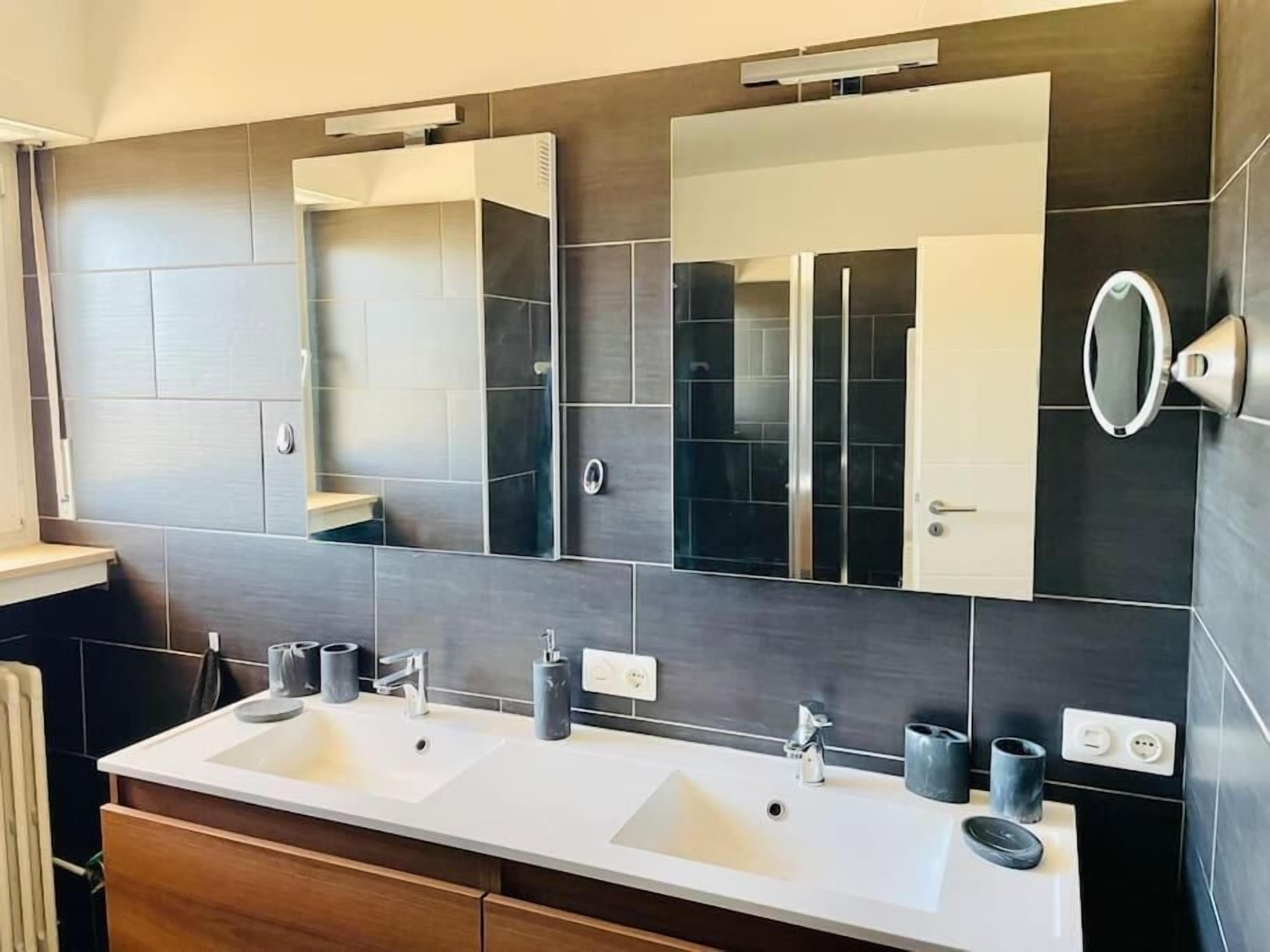 Une salle de bain moderne avec deux lavabos, un grand miroir et des murs recouverts de carreaux foncés.