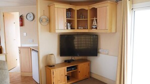 A living room with a flat-screen TV, wooden cabinets, a small kitchen area, and a dining table. - Pets go Free Family 3 Bed Caravan With Decking (King's Lynn)