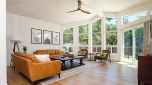 A spacious living room with a large sofa, coffee table, and chairs, large windows offering a view of trees, and a ceiling fan. - Doodlebug de Sedona • Amenity galore (Sedona)