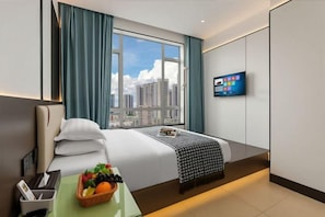 A hotel room with a bed, a TV, a window with a city view, and a fruit basket on the side table. - Dongqi Business Hotel (Dongguan)