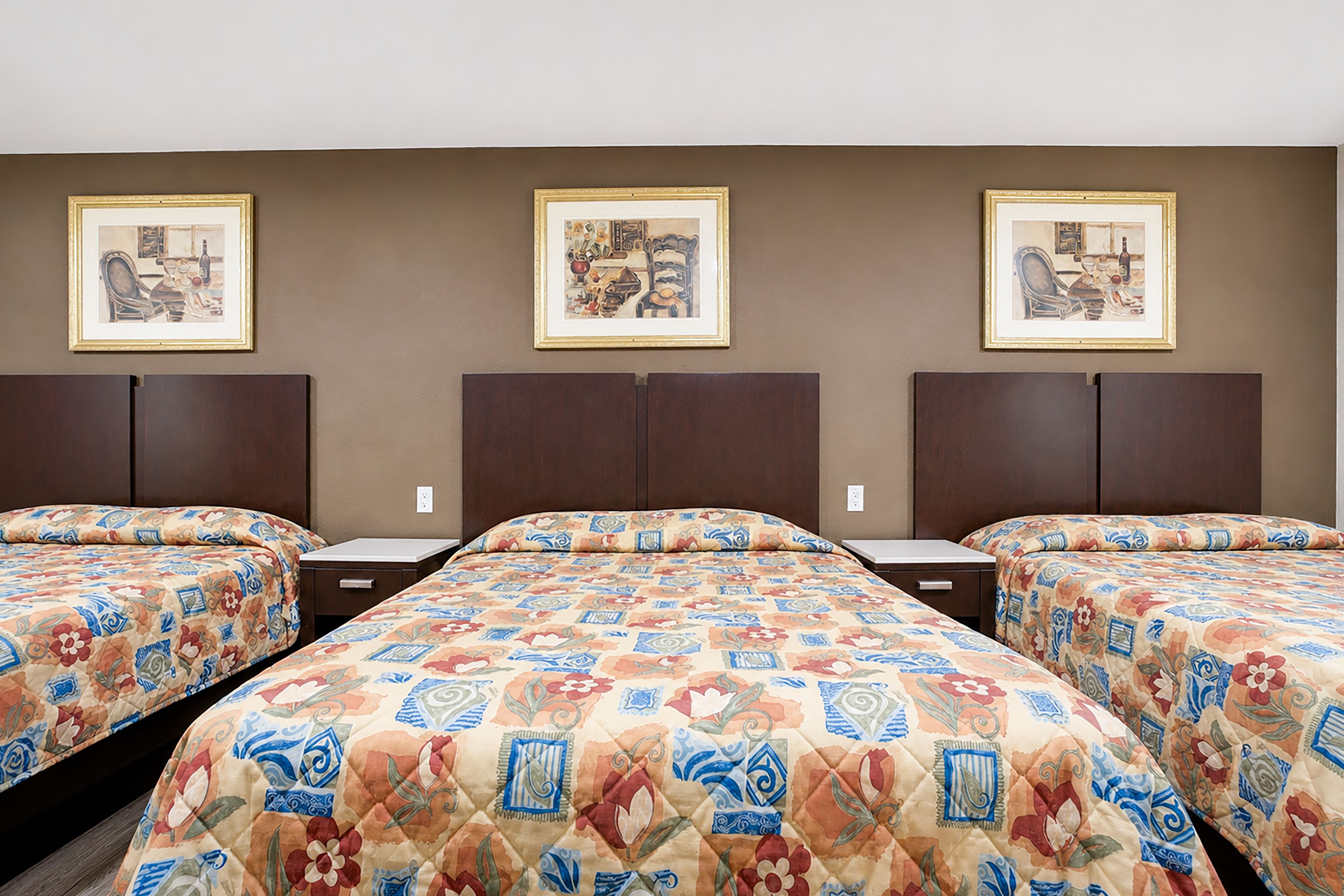 Ein Hotelzimmer mit drei Betten, jedes mit einer gemusterten Tagesdecke, und drei gerahmten Bildern an der Wand.