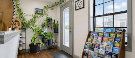 A hallway with wooden flooring, a door, a window with a view, and a wall with potted plants and a travel information stand.