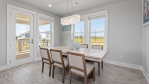 A dining area with a large table and chairs, a chandelier, and large windows offering a view of the outdoors. - Beach View 4BR Retreat | Soccer Field | Putt-Putt | Pet Friendly (Galveston)