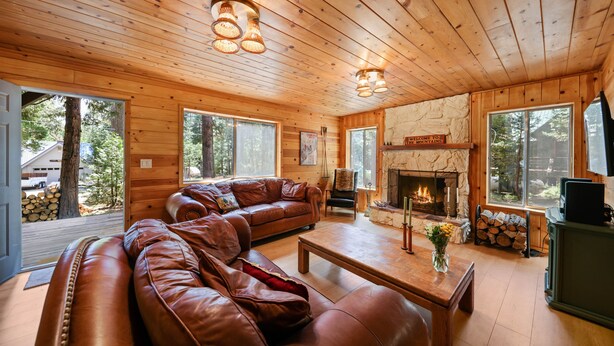 A cozy cabin interior with a leather sectional sofa, a fireplace, and large windows offering views of the surrounding forest. - The Wildwood Cabin - Tahoe National Forest (Emigrant Gap)
