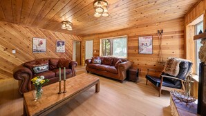 A wooden cabin interior with a wooden ceiling, two leather sofas, a wooden coffee table, and a window with a view of trees. - The Wildwood Cabin - Tahoe National Forest (Emigrant Gap)
