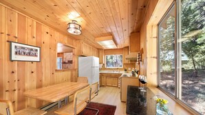 A wooden interior with a dining table, chairs, a refrigerator, and a large window offering a view of trees. - The Wildwood Cabin - Tahoe National Forest (Emigrant Gap)