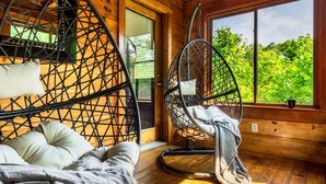 A cozy room with a wooden interior, a bed with pillows, a hanging chair with a cushion, and a window with a view of greenery. - Private*Pool*HotTub*Sauna*FirePit*Theater+GameRoom (Sevierville)