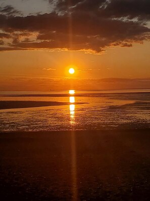 Un atardecer sobre una playa, con el sol bajo en el horizonte, proyectando un reflejo en el agua.