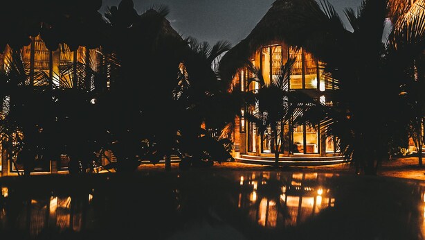 Un resort con varias construcciones, palmeras y una piscina reflectante de noche.