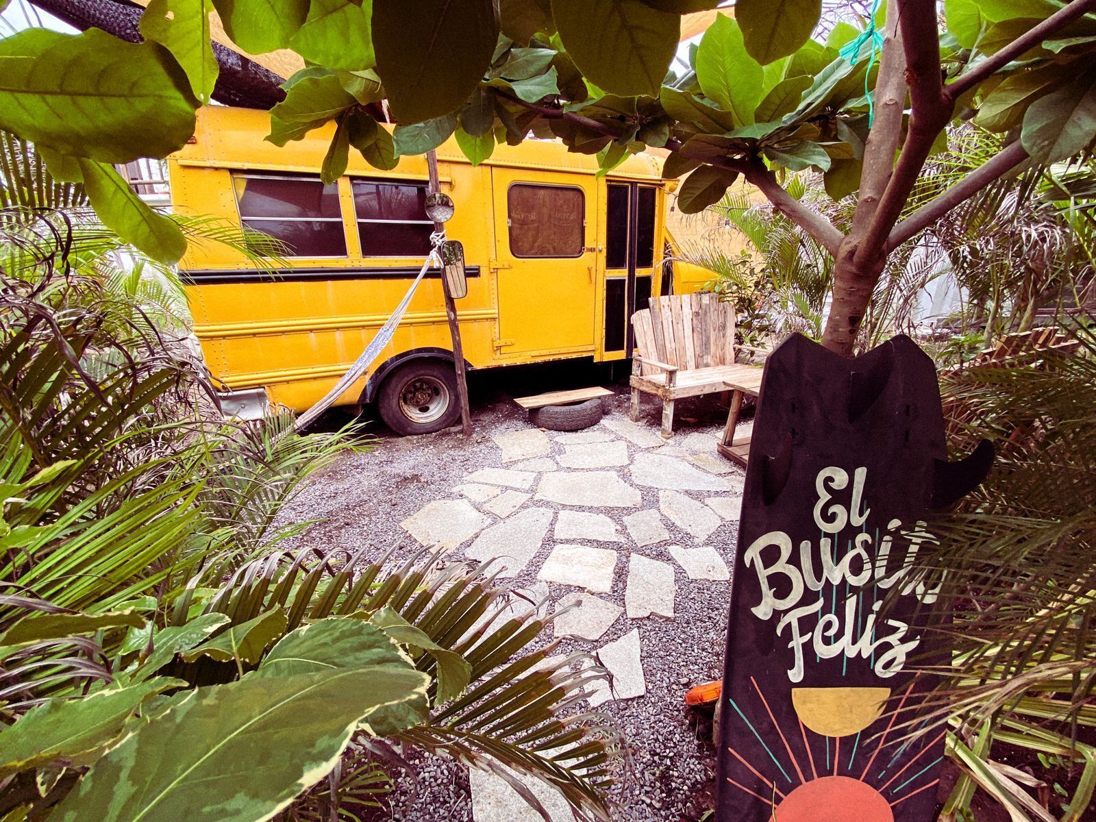 Un autobús amarillo estacionado en un jardín con un camino de piedra, una silla de madera y un letrero que dice "El Bus del Feliz".