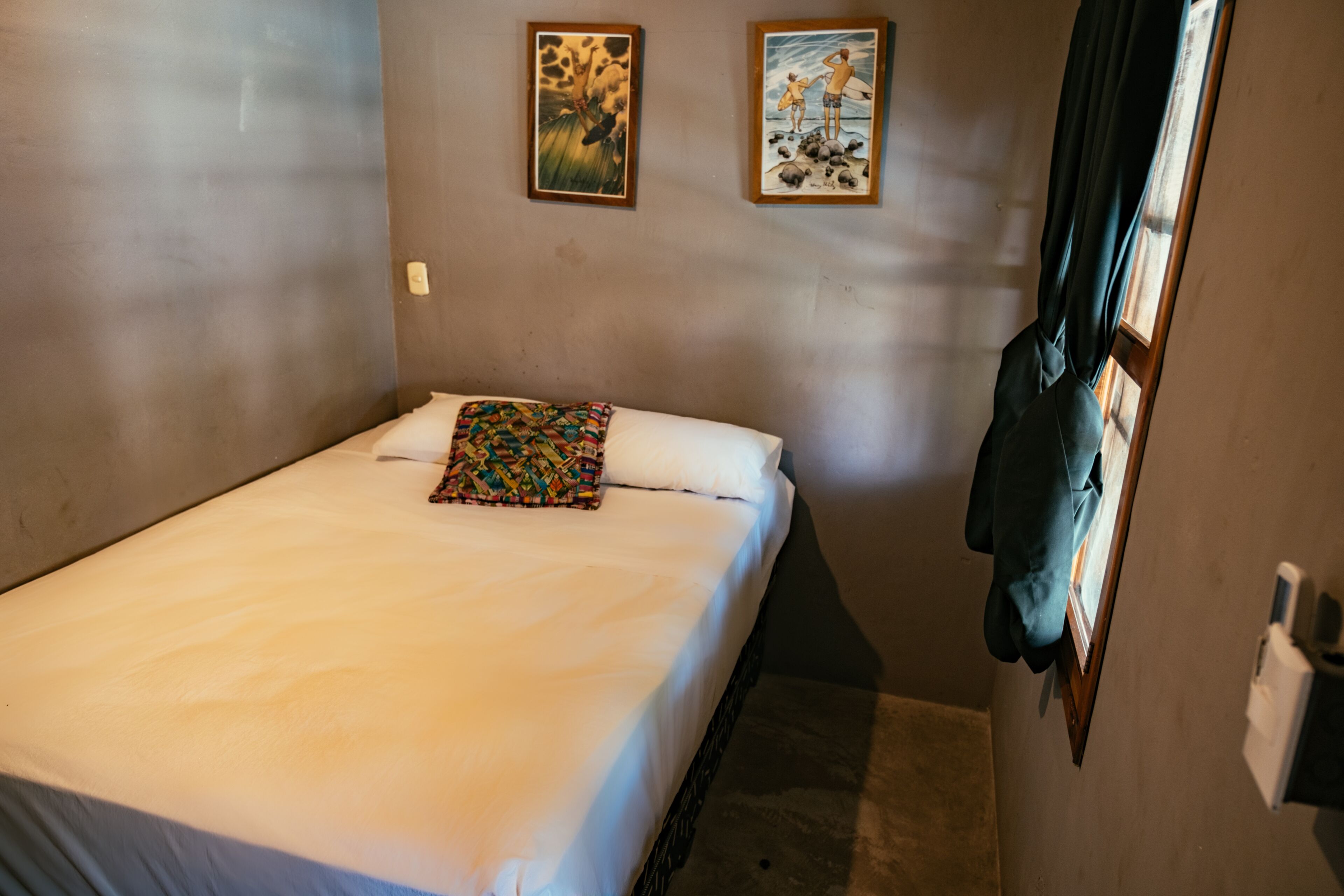Une petite chambre avec un lit simple, un mur uni, un coussin coloré et deux tableaux encadrés au-dessus du lit.