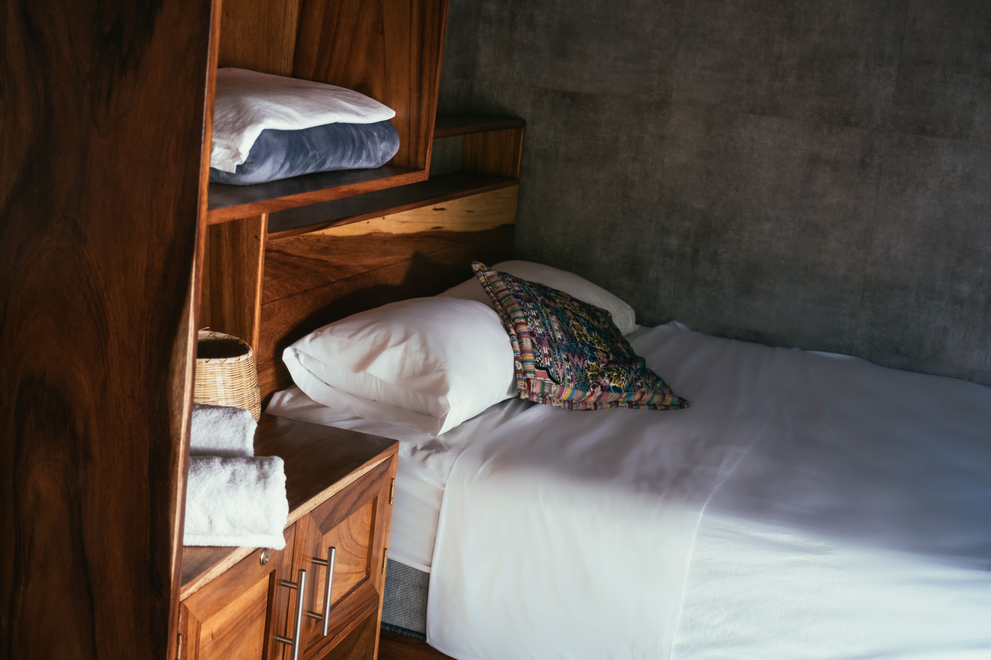 Ein Schlafzimmer mit einem Bett, einem Nachttisch aus Holz, einem Regal mit Kissen und einer strukturierten Wand.