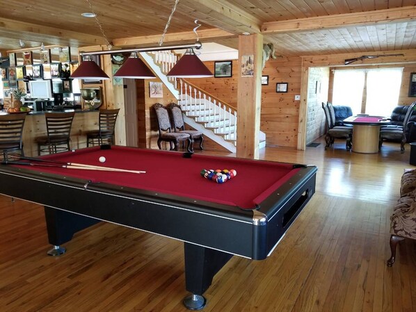 Una mesa de billar en una sala con suelo de madera, una escalera y un área de descanso.