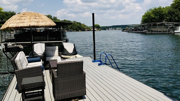 Une terrasse en bois, agrémentée de meubles en osier et d’un parasol en chaume, offre une vue sur un lac paisible, avec un quai et des bâtiments visibles au loin.