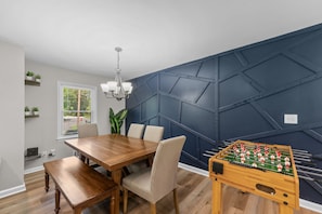 A dining area with a wooden table, chairs, and a foosball table against a dark blue wall with geometric patterns.