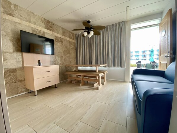 A modern living room with a flat-screen TV, wooden furniture, a ceiling fan, and a view of the outside. - The VIP Family Motel (Wildwood Crest)
