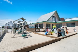 A playground with swings and slides, a wooden deck, and a building with a green roof and windows. - The VIP Family Motel (Wildwood Crest)