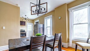 A dining area with a wooden table, chairs, a chandelier, and a view of the city through large windows. - The Parkside by Jungle House – Short North Boutique Suites (Columbus)