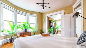 A bedroom with a large bed, multiple potted plants, a vintage trunk, and a chandelier. - The Parkside by Jungle House – Short North Boutique Suites (Columbus)