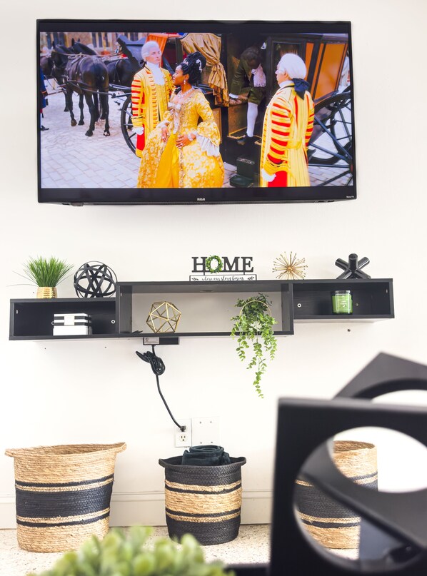 A television mounted on a wall displaying a scene with people in historical attire and horses. Below, a shelf holds decorative items and a power cord.