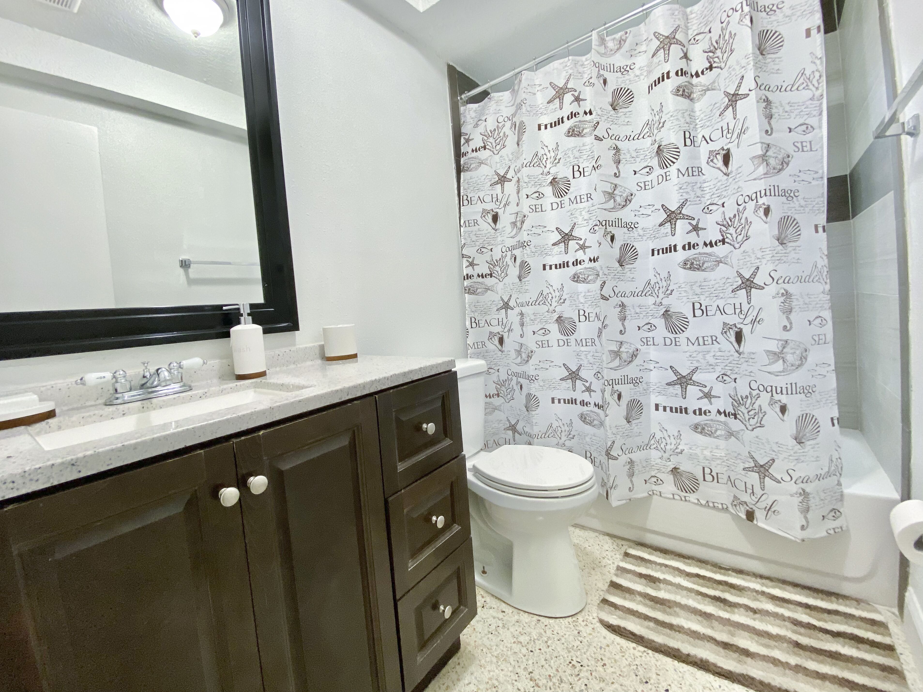 Un baño con lavamanos blanco, muebles de madera oscura, inodoro blanco y cortina de ducha con motivos playeros.