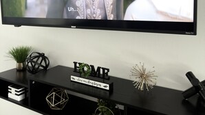 Un téléviseur fixé au mur affiche une scène mettant en scène deux personnages. En dessous du téléviseur, on trouve une étagère noire ornée d’objets décoratifs et d’une pancarte sur laquelle on peut lire : « CHEZ NOUS, C’EST ICI QUE NOTRE HISTOIRE COMMENCE ».