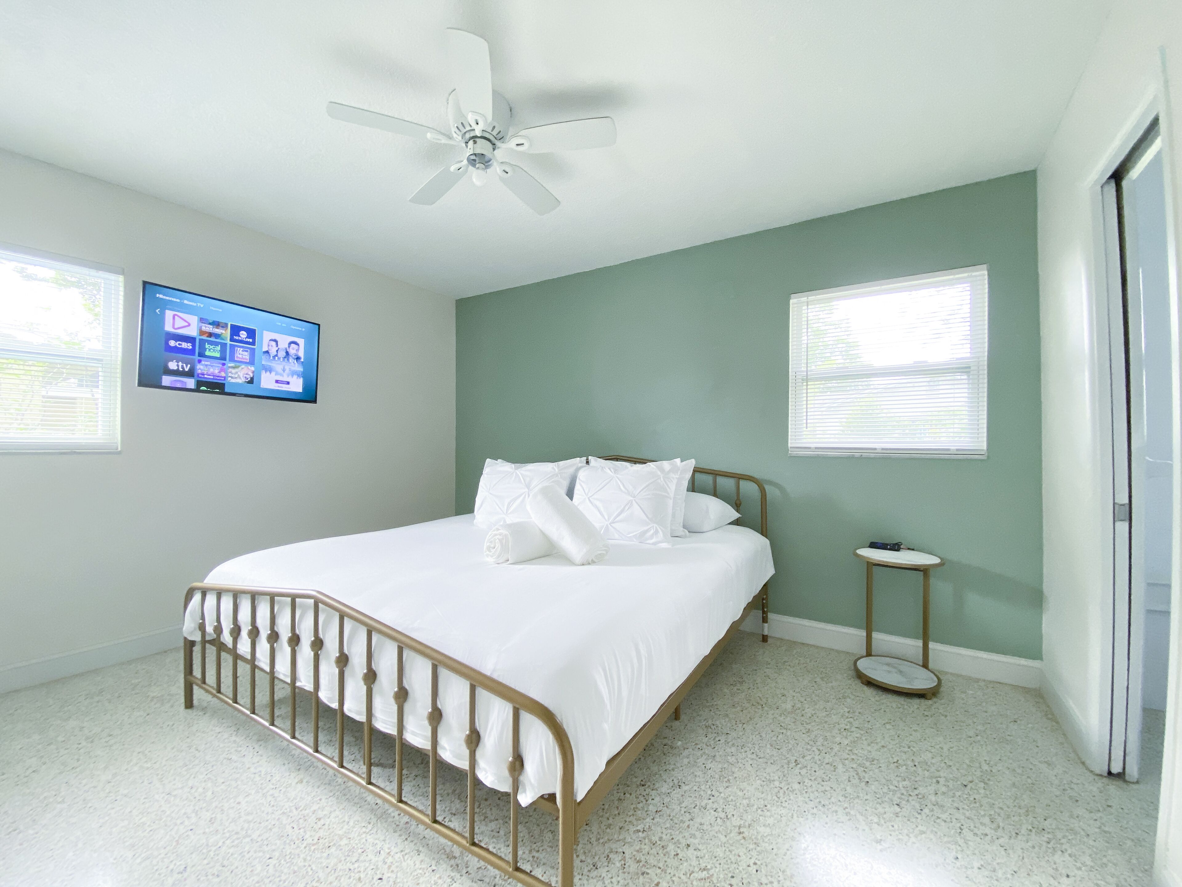 Ein Schlafzimmer mit einem Bett, einem Deckenventilator, einem an der Wand montierten Fernseher und zwei Fenstern.