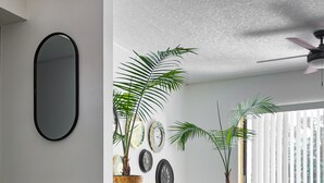 Un salon moderne avec un canapé en cuir, une table basse et des objets décoratifs. On y trouve deux plantes en pot, une horloge murale et un ventilateur de plafond.