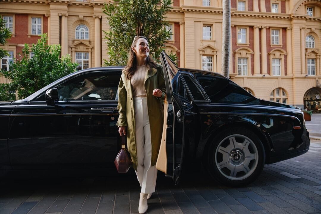 Une femme sort d’une voiture de luxe noire garée devant un bâtiment historique à l’architecture classique.