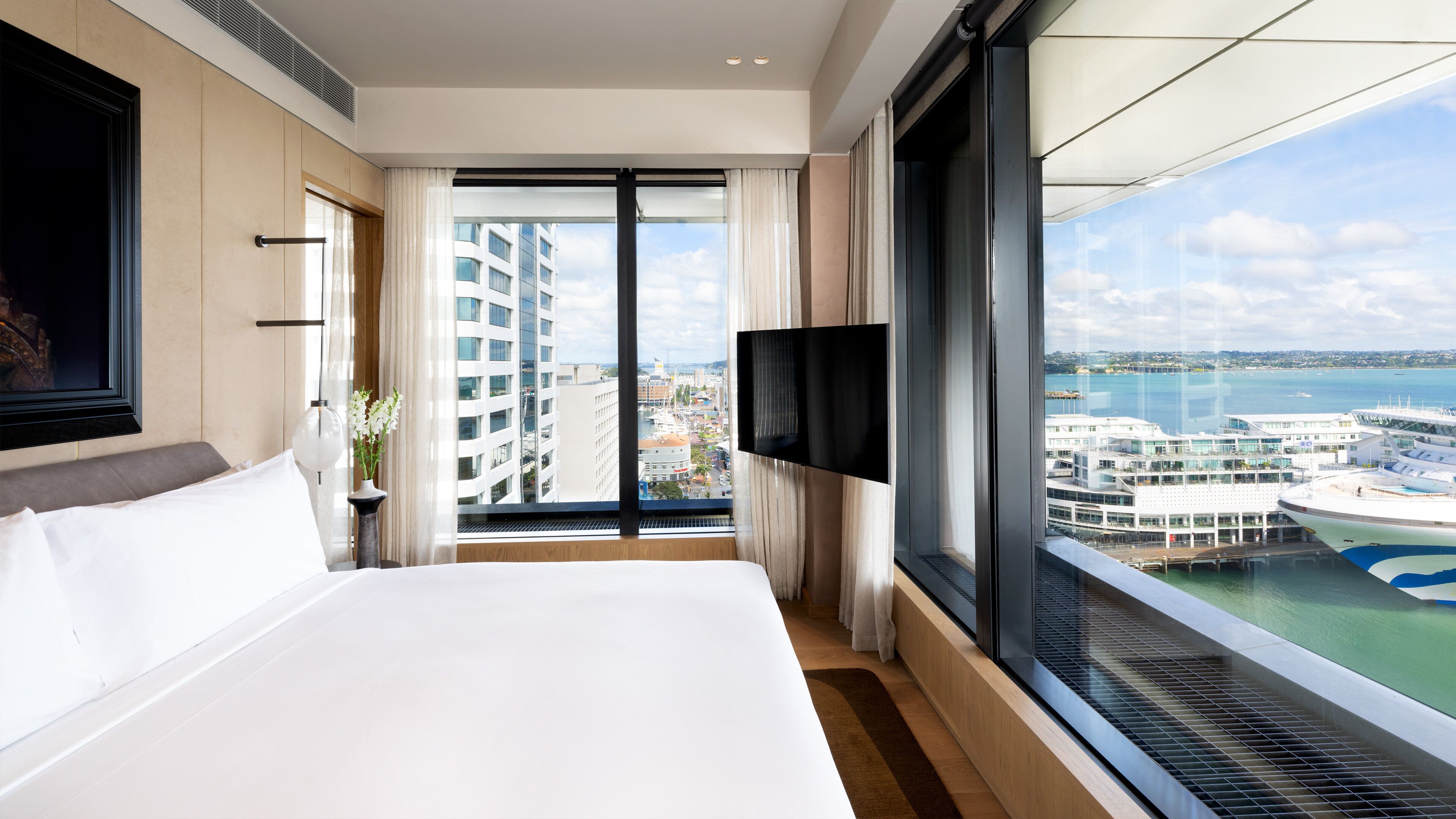 A hotel room with a large bed, a TV mounted on the wall, and a view of a cruise ship and cityscape through a large window.