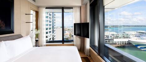 A hotel room with a large bed, a TV mounted on the wall, and a view of a cruise ship and cityscape through a large window.