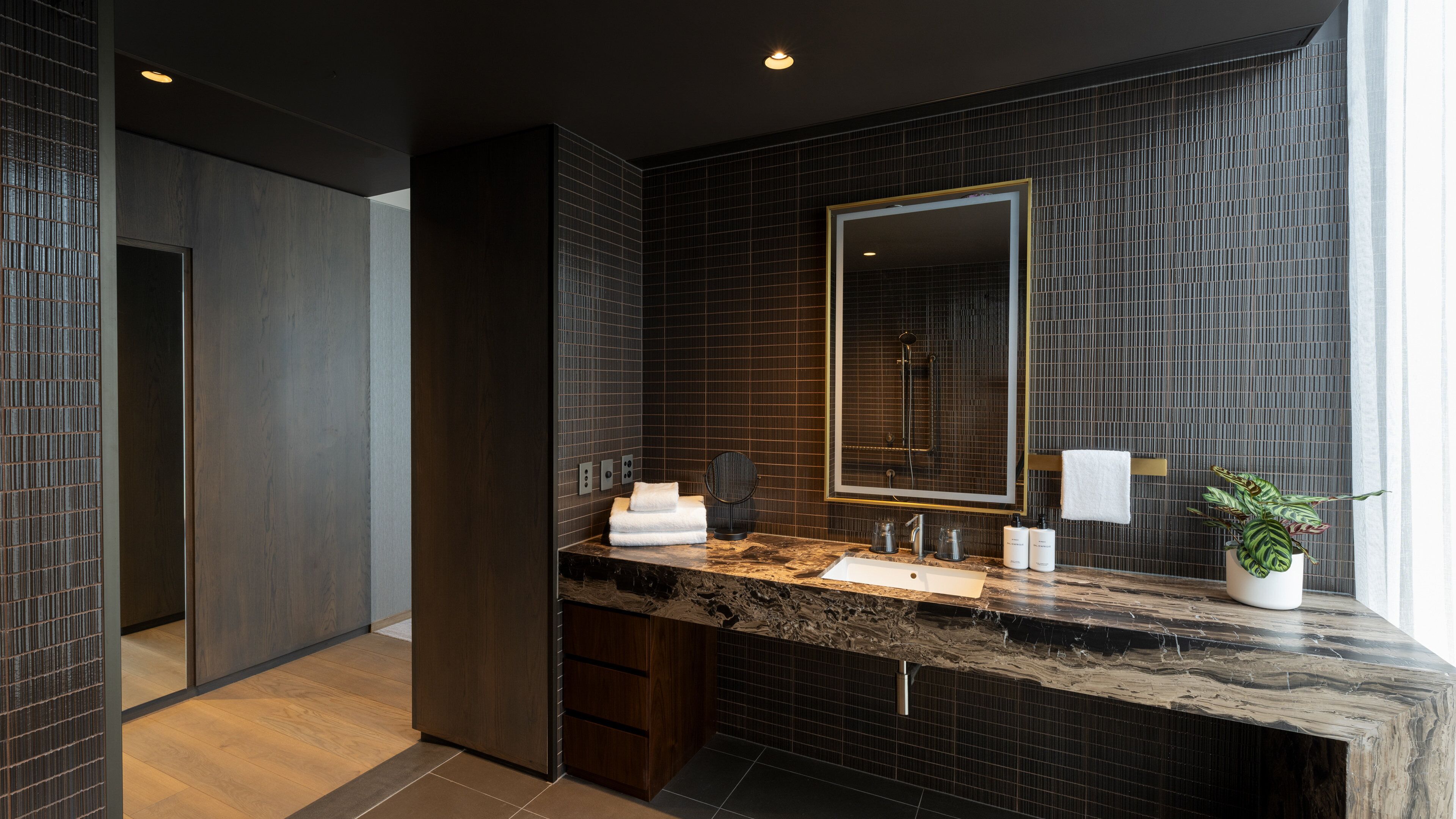 A modern bathroom with a dark tiled wall, a large mirror, and a marble countertop.