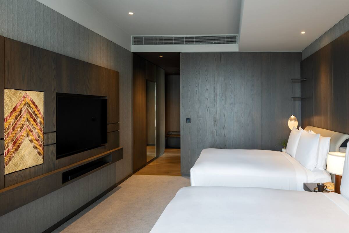 Ein modernes Hotelzimmer mit großem Flachbildfernseher an der Wand, einem Bett mit weißer Bettwäsche und einem Nachttisch mit Lampe.