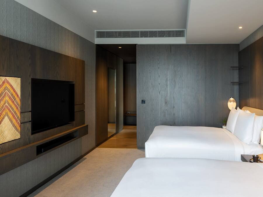 A modern hotel room with a large flat-screen TV mounted on the wall, a bed with white linens, and a bedside table with a lamp.