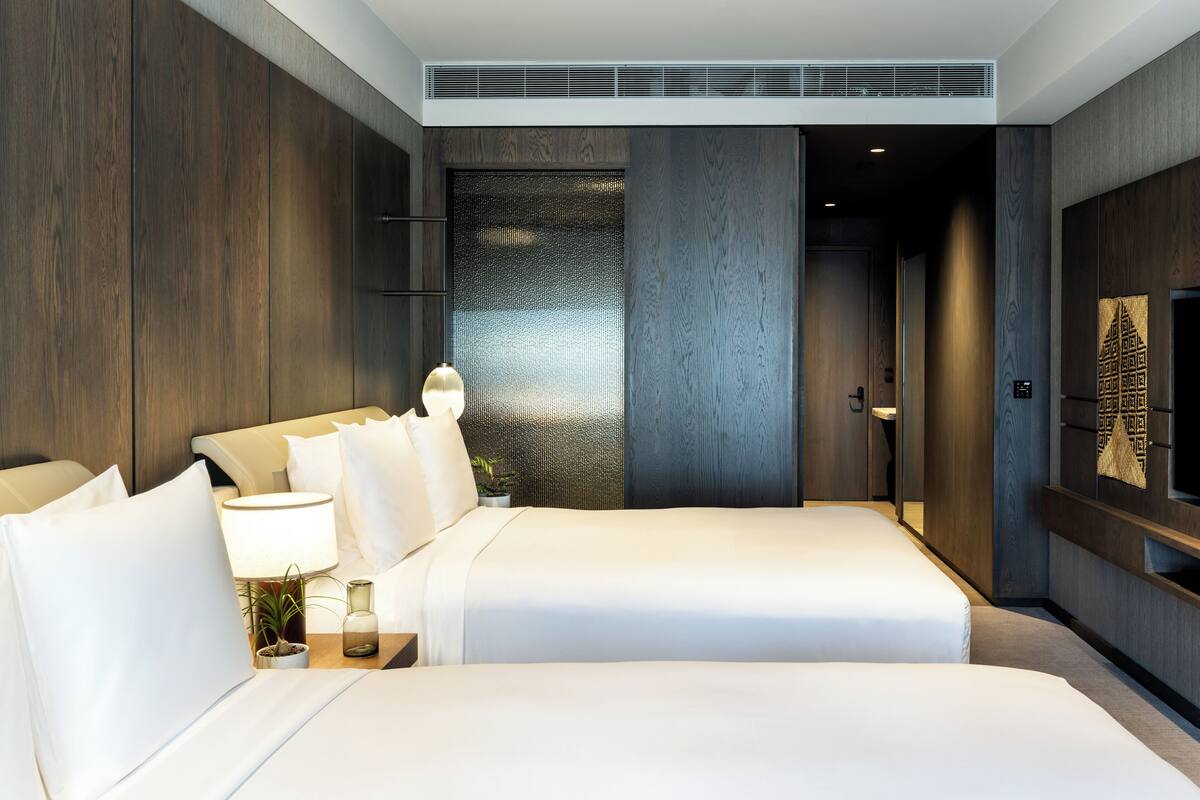 Ein modernes Hotelzimmer mit zwei Betten, einem großen Spiegel, einem wandmontierten Fernseher und einem kleinen Tisch mit einer Lampe und einer Pflanze.