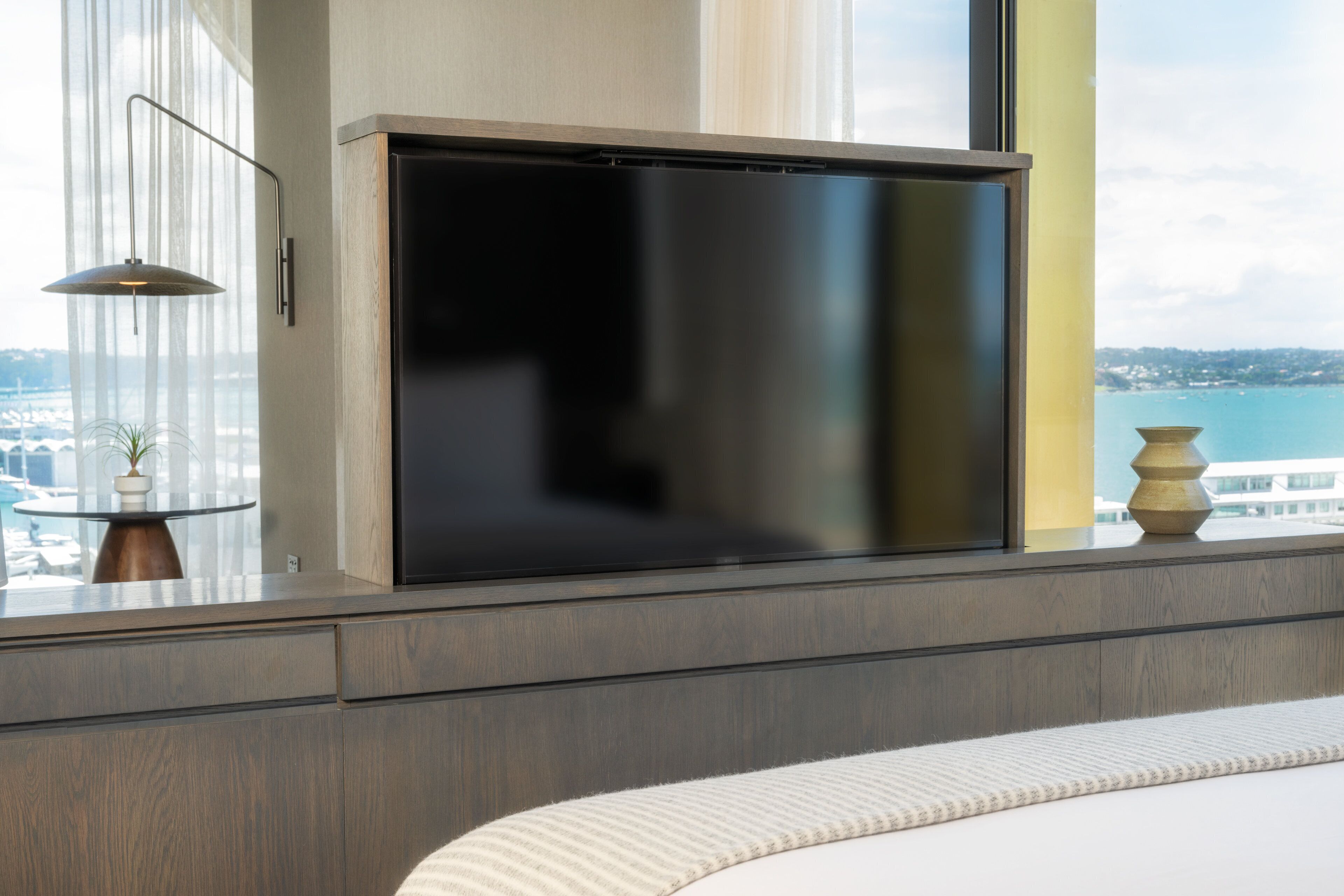 Habitación de hotel moderna con un televisor de pantalla plana grande montado en un mueble de madera, una mesa redonda de cristal con una planta en maceta y vistas al mar a través de amplios ventanales.