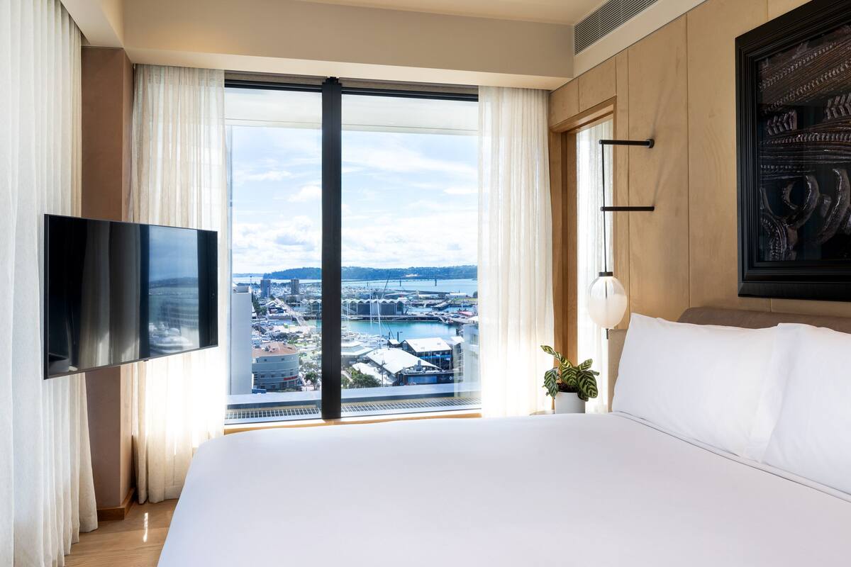 Ein Hotelzimmer mit einem großen Bett, einem flachbildfernseher an der Wand, Blick auf eine Stadtlandschaft durch ein großes Fenster und einem dekorativen Wandbild.